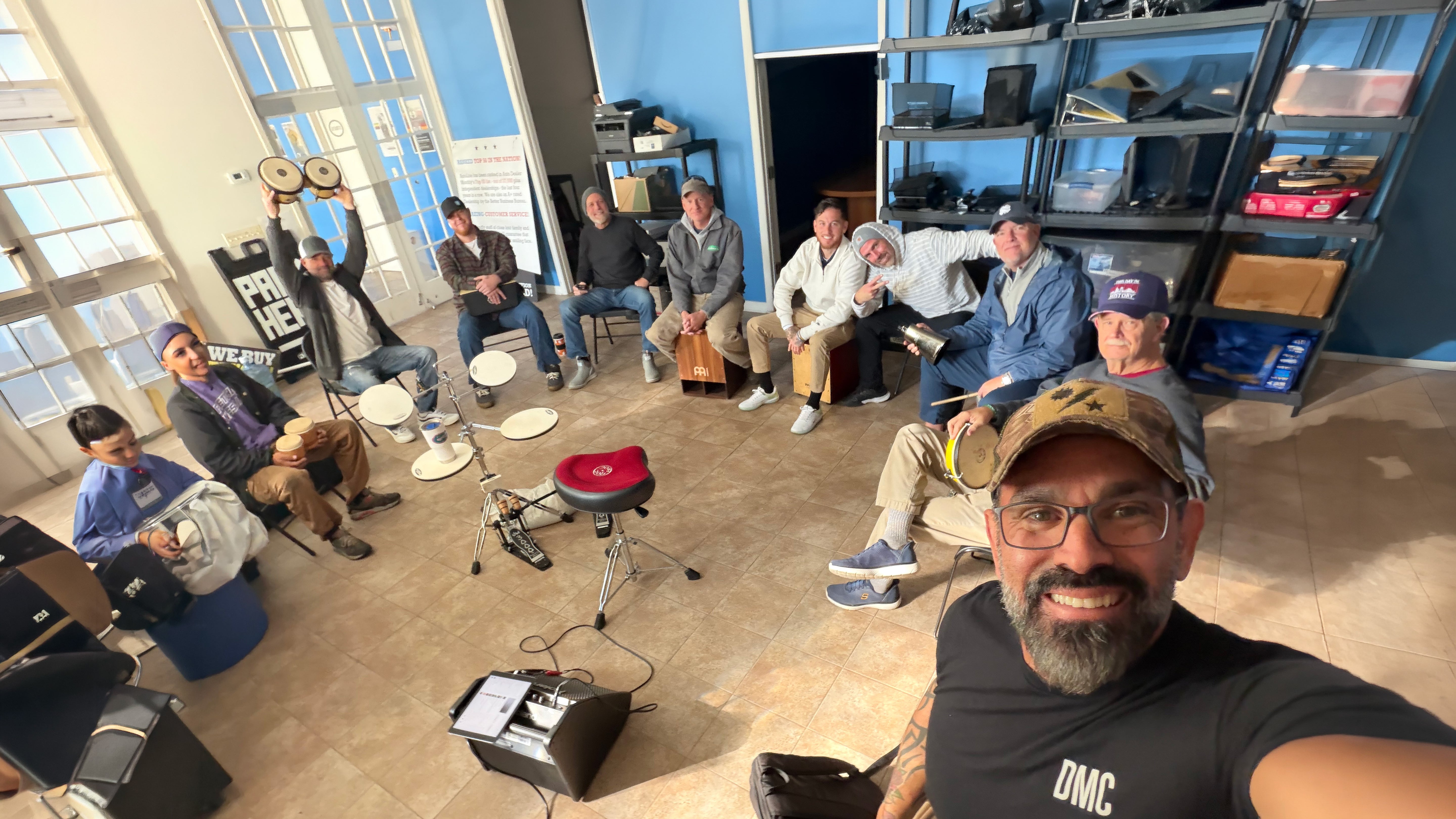 A group of people gathered in a studio or workshop space with blue walls, shelves, windows, and musical equipment, including drums, a red stool, and a speaker. One man in the foreground is taking a selfie while others sit and stand in a circle-like arrangement.