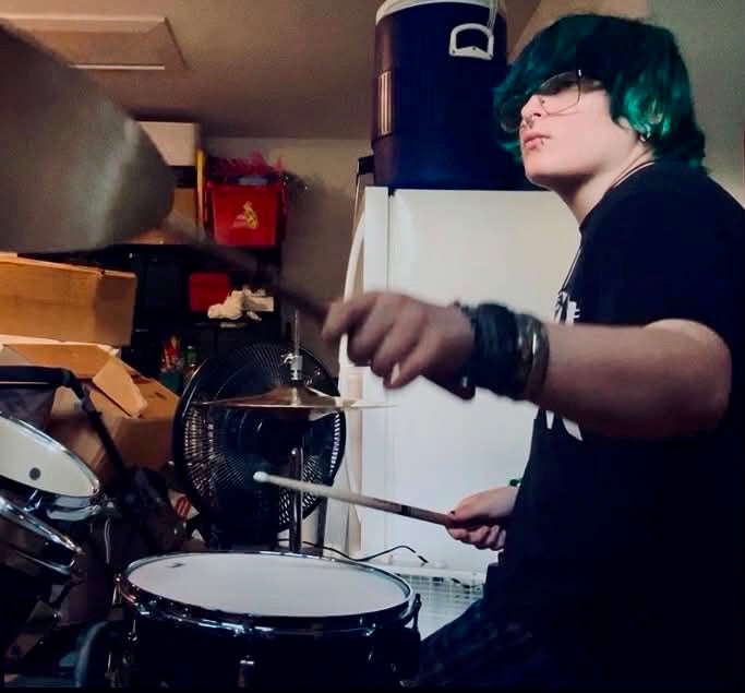A person with bright green hair and glasses playing a drum kit in a cluttered indoor room, wearing a black t-shirt and multiple bracelets.