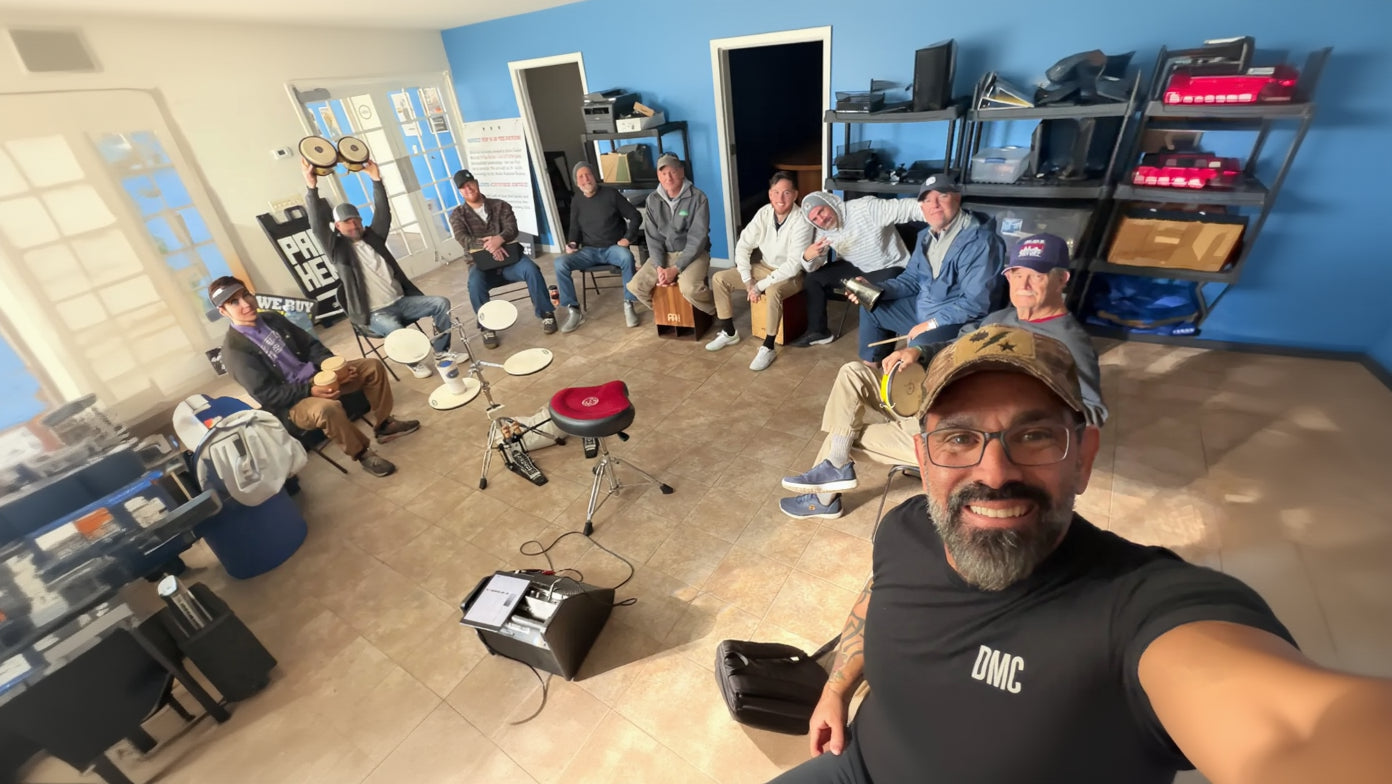 A group of men gathered indoors in a workshop or studio space, sitting in a circle with hand drums, a red stool, a speaker, and shelves in the background. One man in the foreground is taking a selfie wearing a cap and glasses, and another man holds a drum overhead in the upper left.