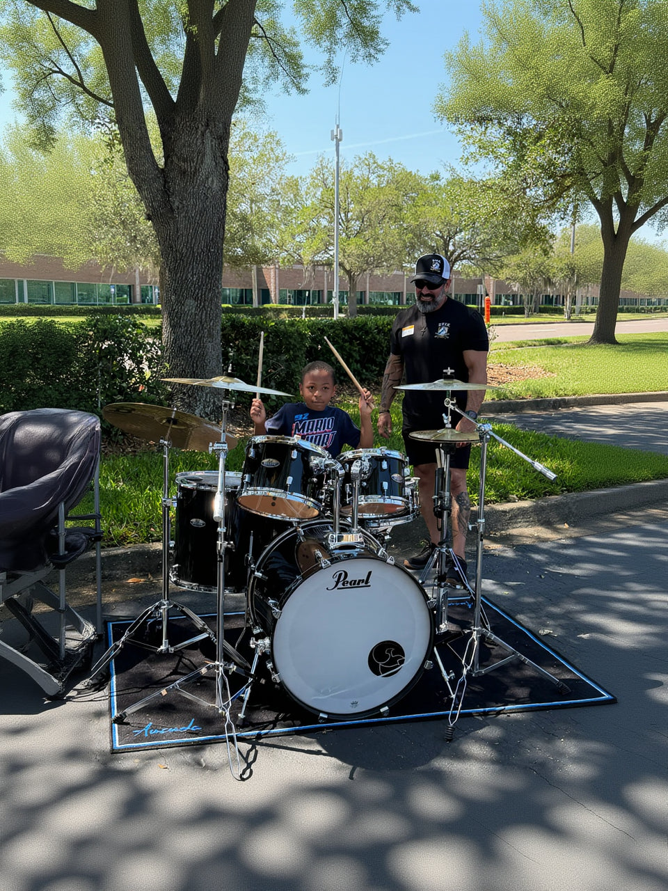 A young boy sits behind a black Pearl drum kit while a man stands beside him outdoors on a paved area with trees and grass in the background.
