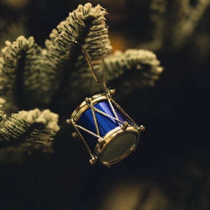 A miniature blue and gold drum Christmas ornament hanging from a snowy pine tree branch.