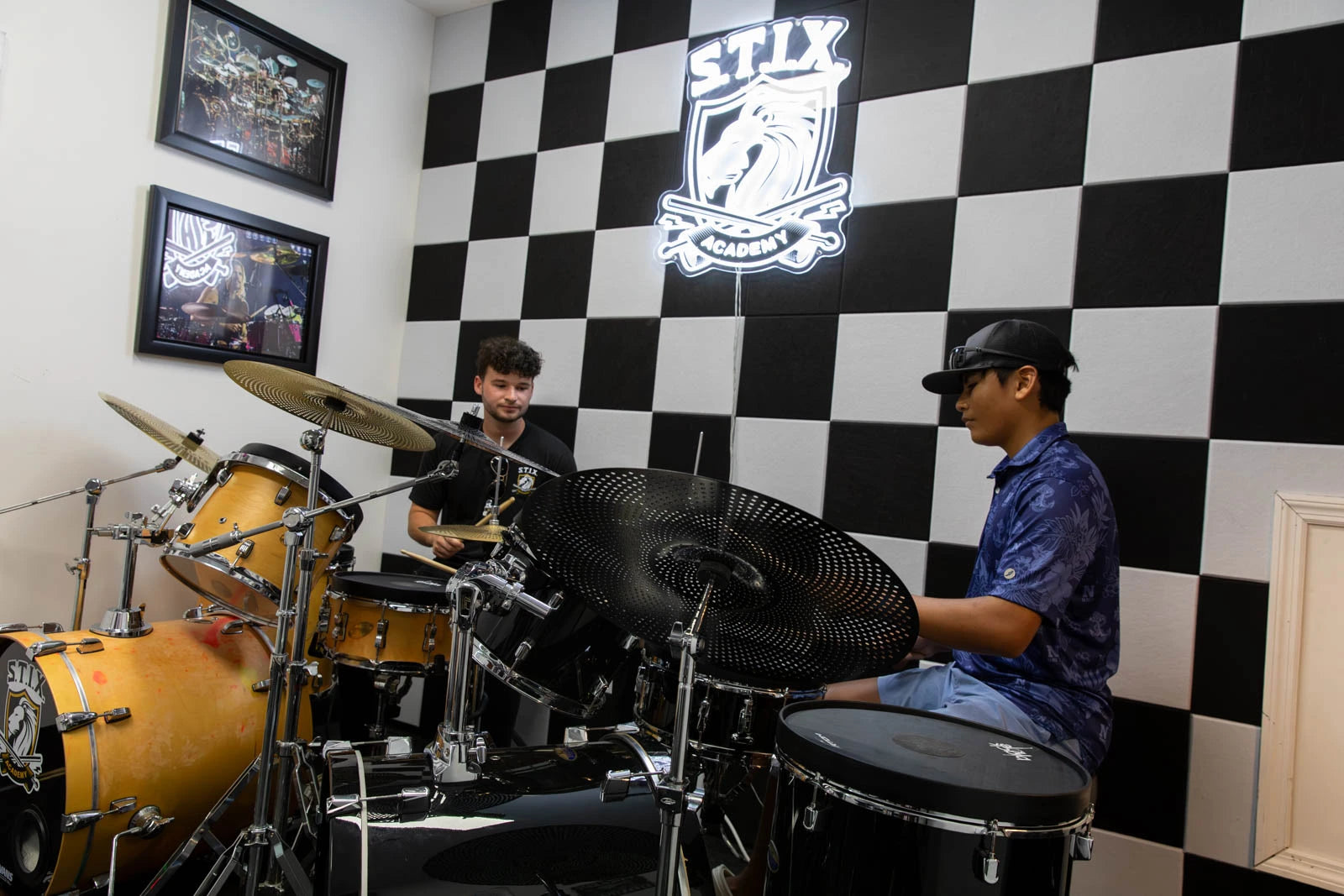 A drummer wearing a black cap and blue patterned shirt sits behind a black drum kit in a room with a black and white checkered wall. Another person is visible behind the kit.