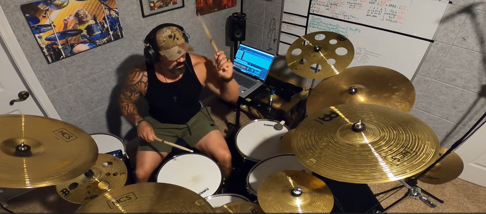 A man wearing a camouflage cap, black tank top, and green shorts sits behind a full drum kit with multiple gold cymbals, playing with drumsticks in a room with a whiteboard and posters on the wall.