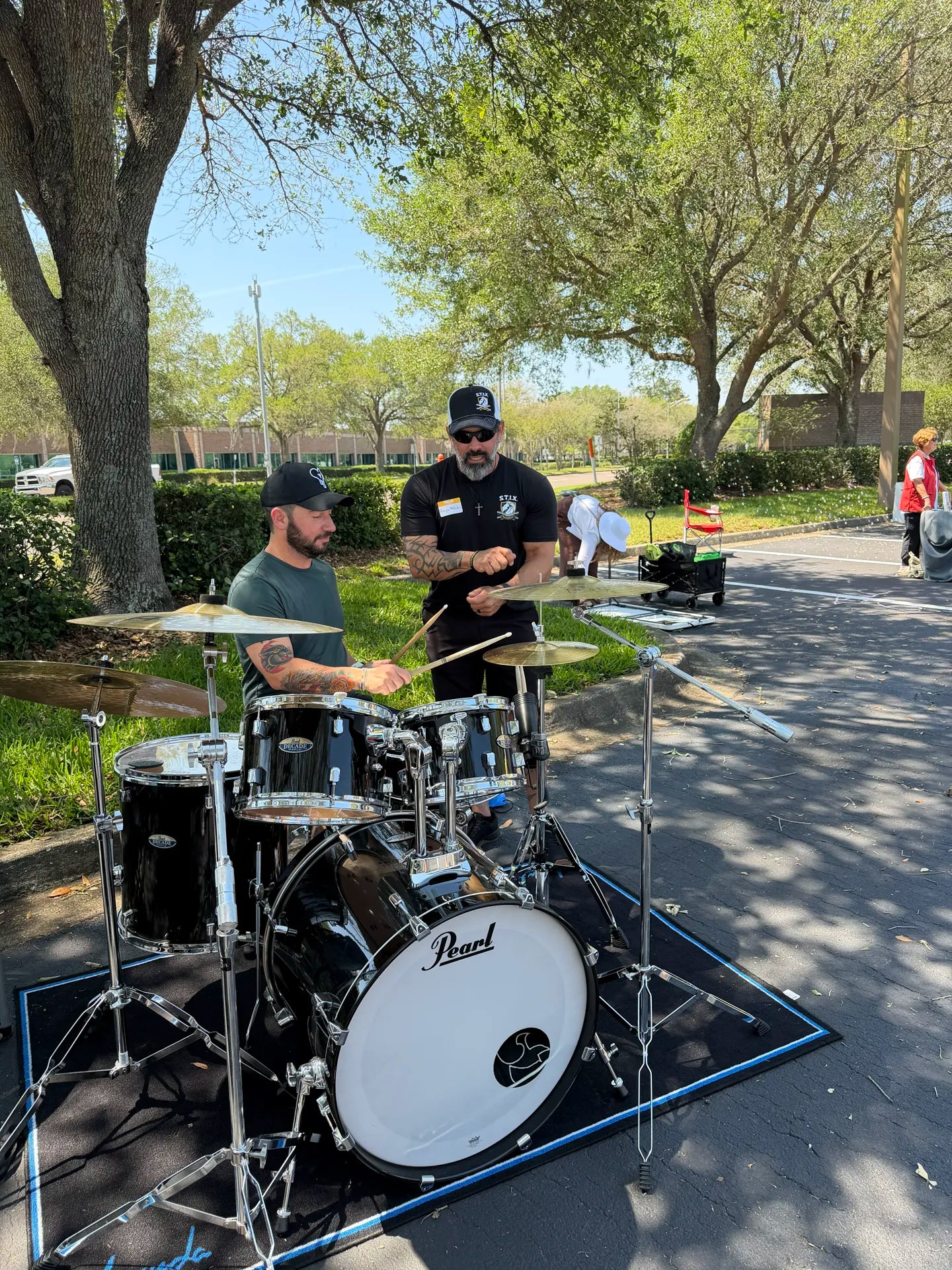 A man stands behind a black Pearl drum kit while another man sits at the drum set in an outdoor park setting with trees and a paved path.
