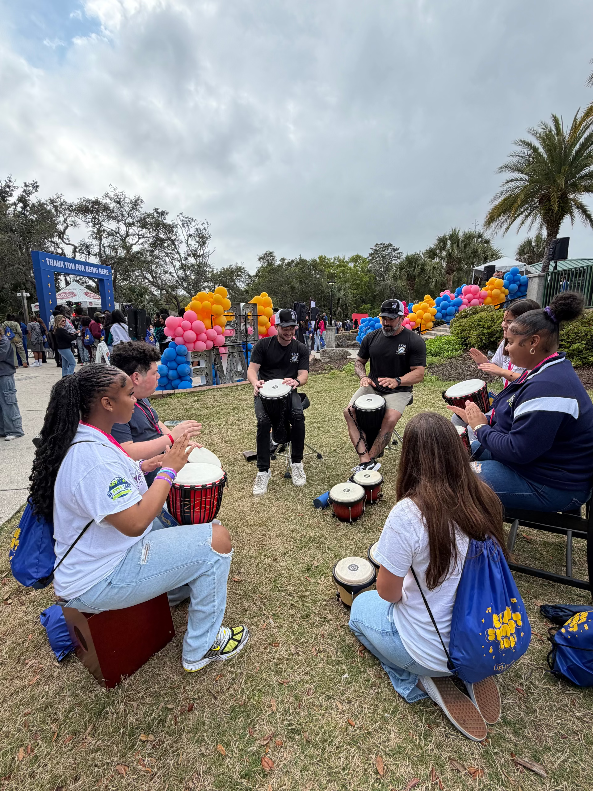 A group of people outdoors on grass playing hand drums, with balloons, trees, and a cloudy sky in the background.