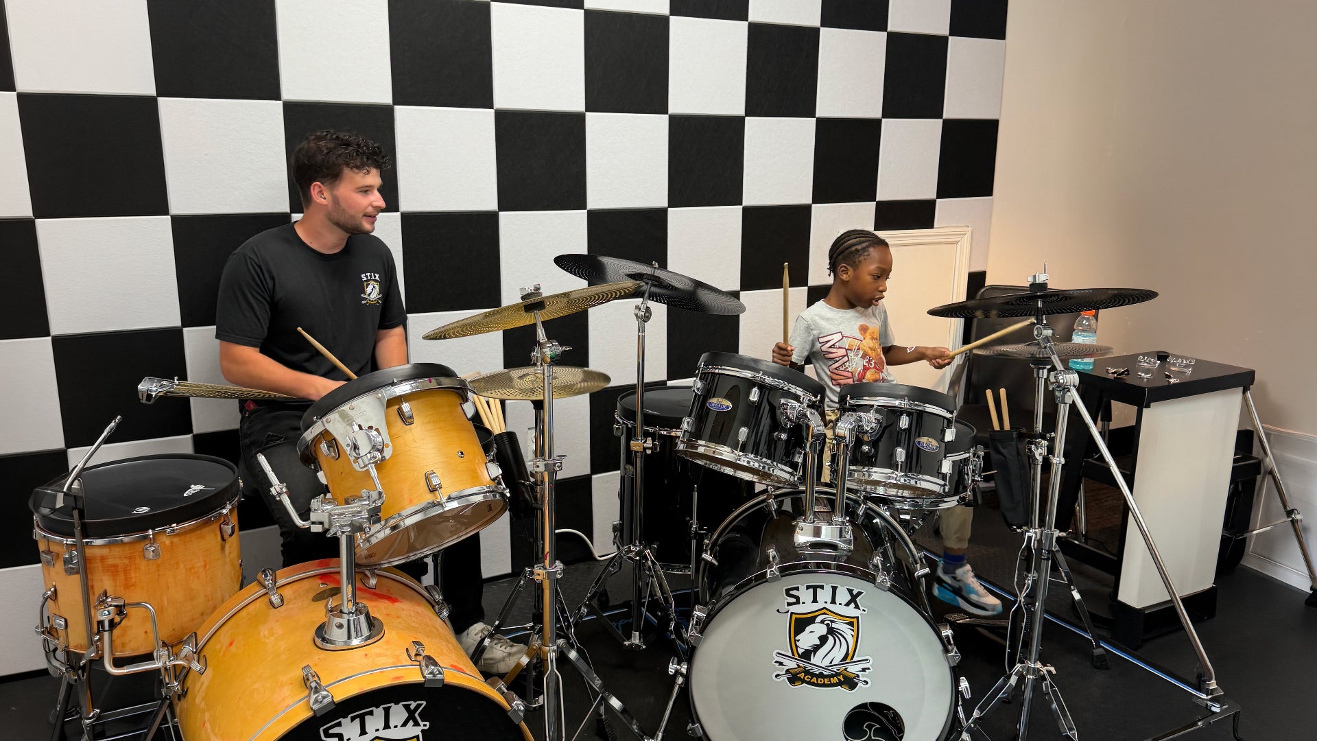 A man and a child playing a STIX branded drum set in a room with a black and white checkered wall.