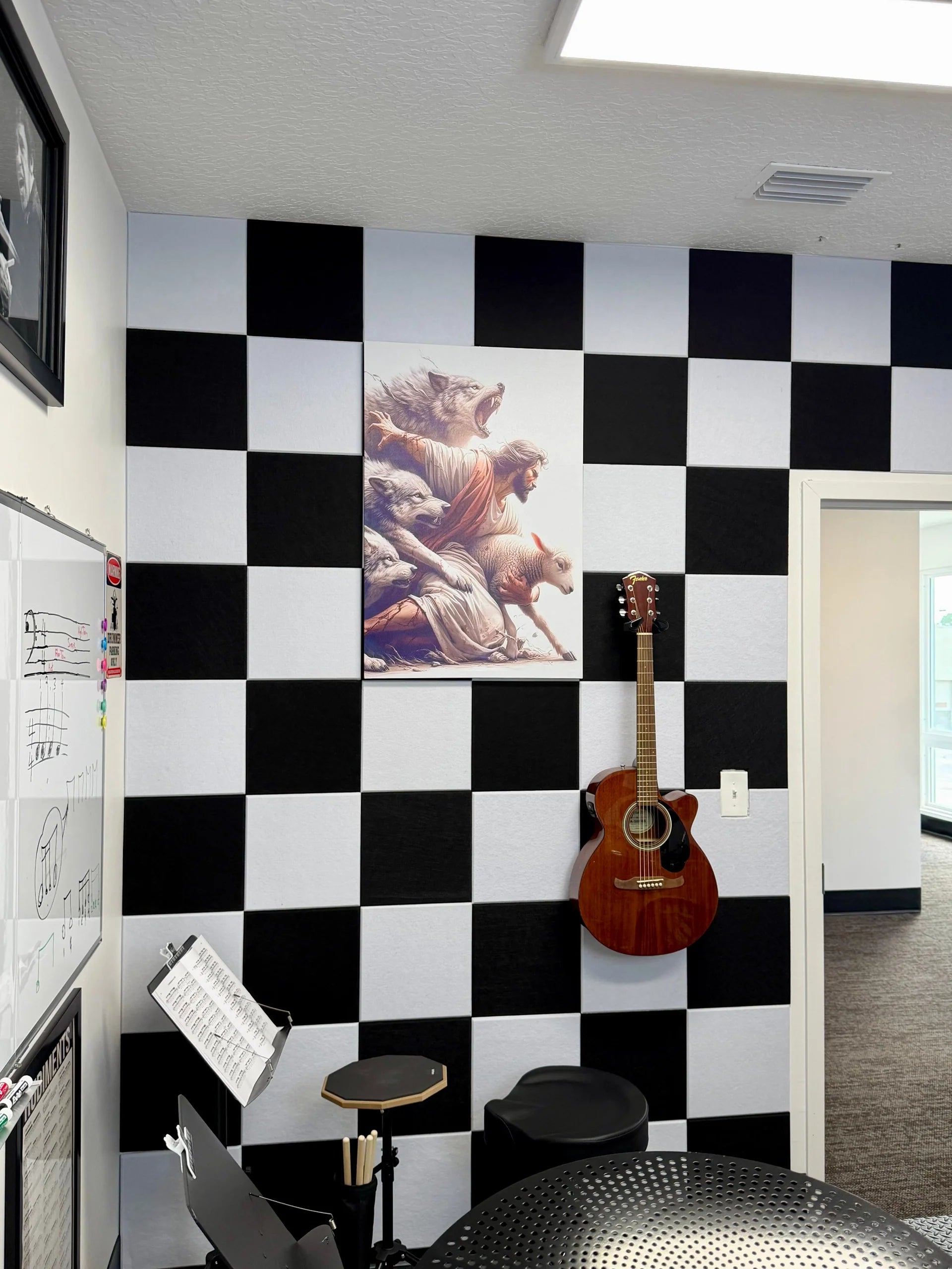 A black and white checkered wall in a room, featuring a framed classical painting of a Jesus, a brown acoustic guitar hanging on the wall, and music stands in the foreground.