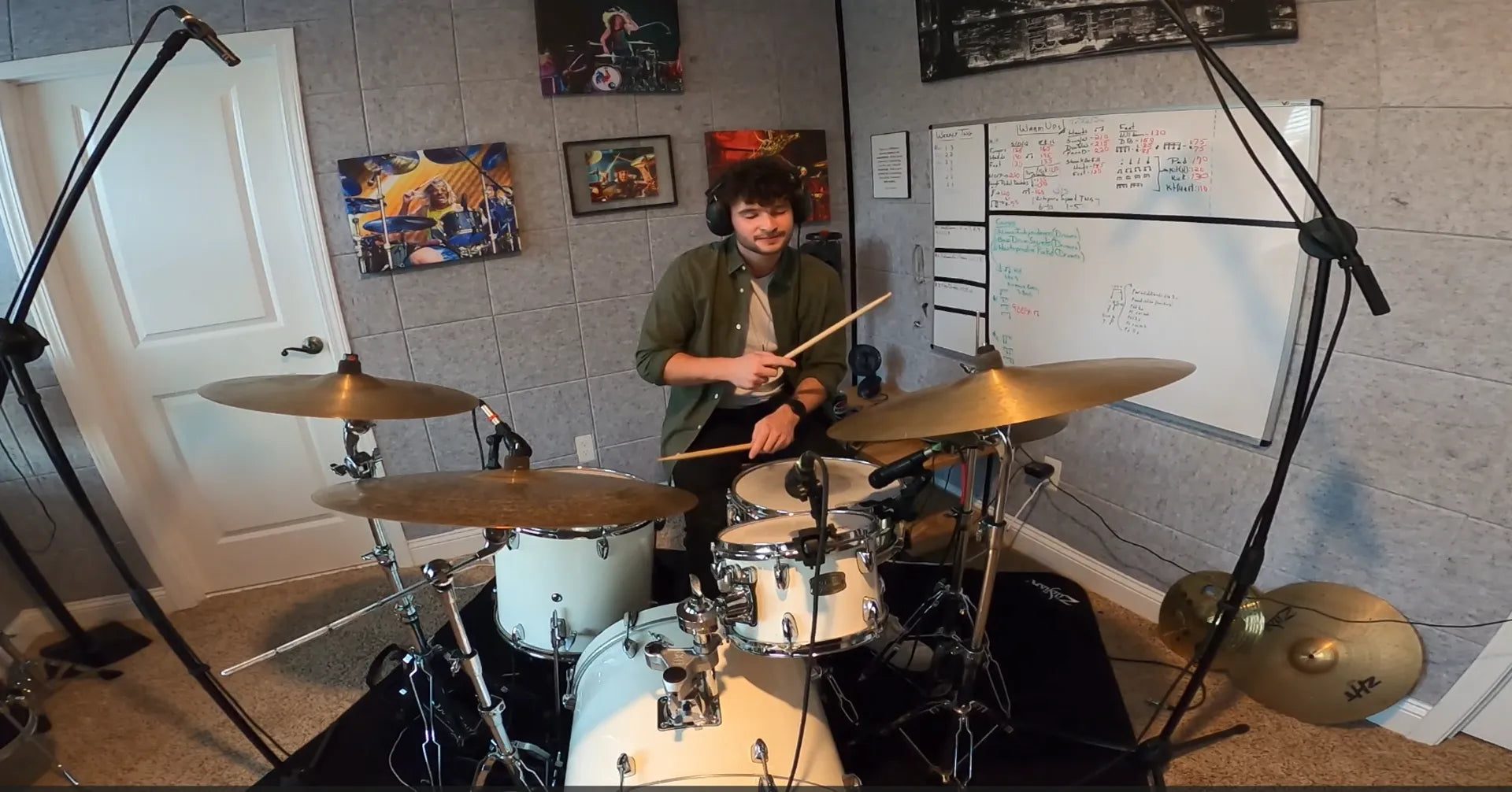 A person standing behind a white drum kit in a room with wall art, a whiteboard, and microphone stands visible around the kit.