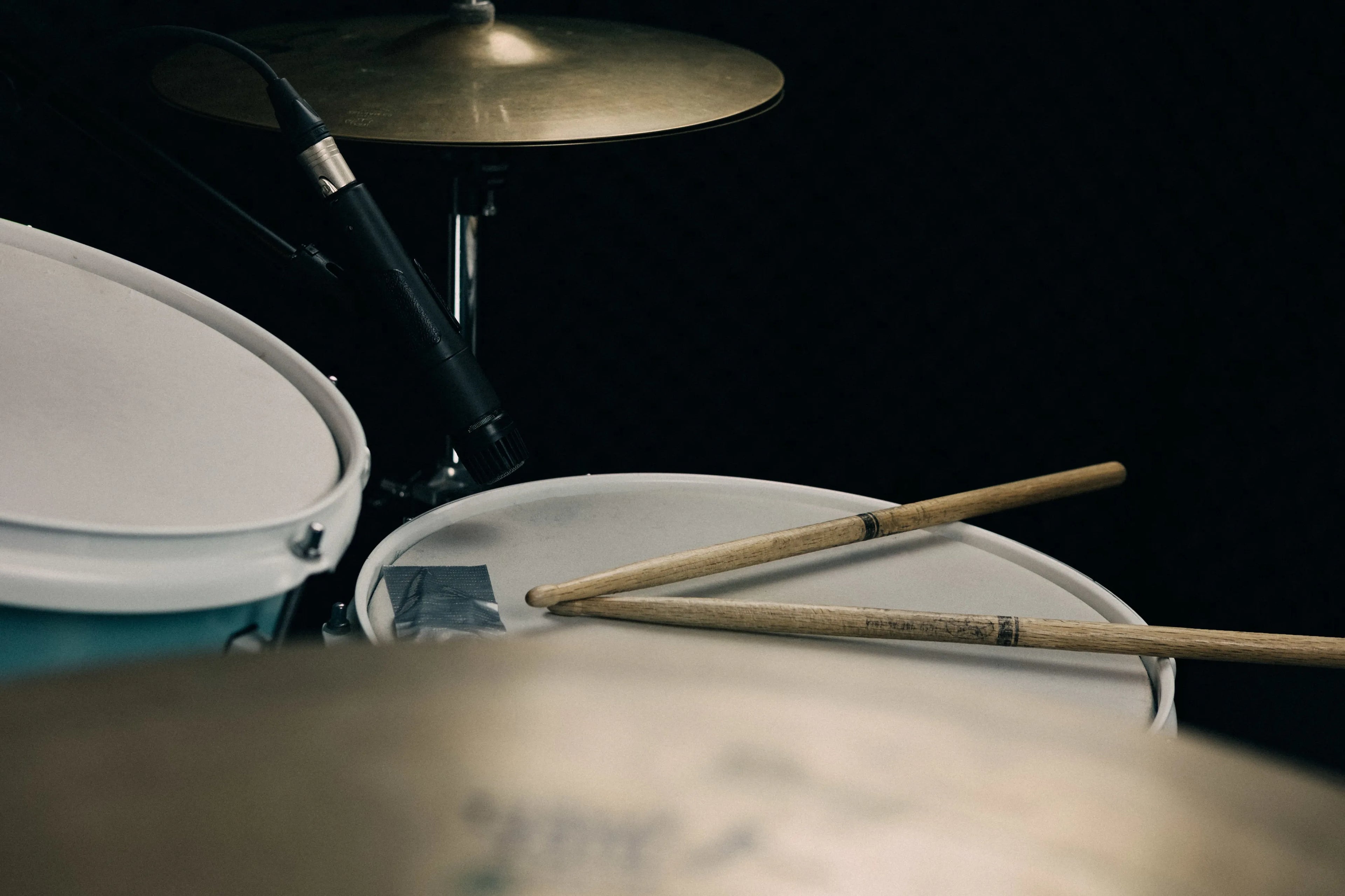 Close-up of a drum set with white drum shells, a brass cymbal, and two wooden drumsticks resting on the drum head against a dark background.