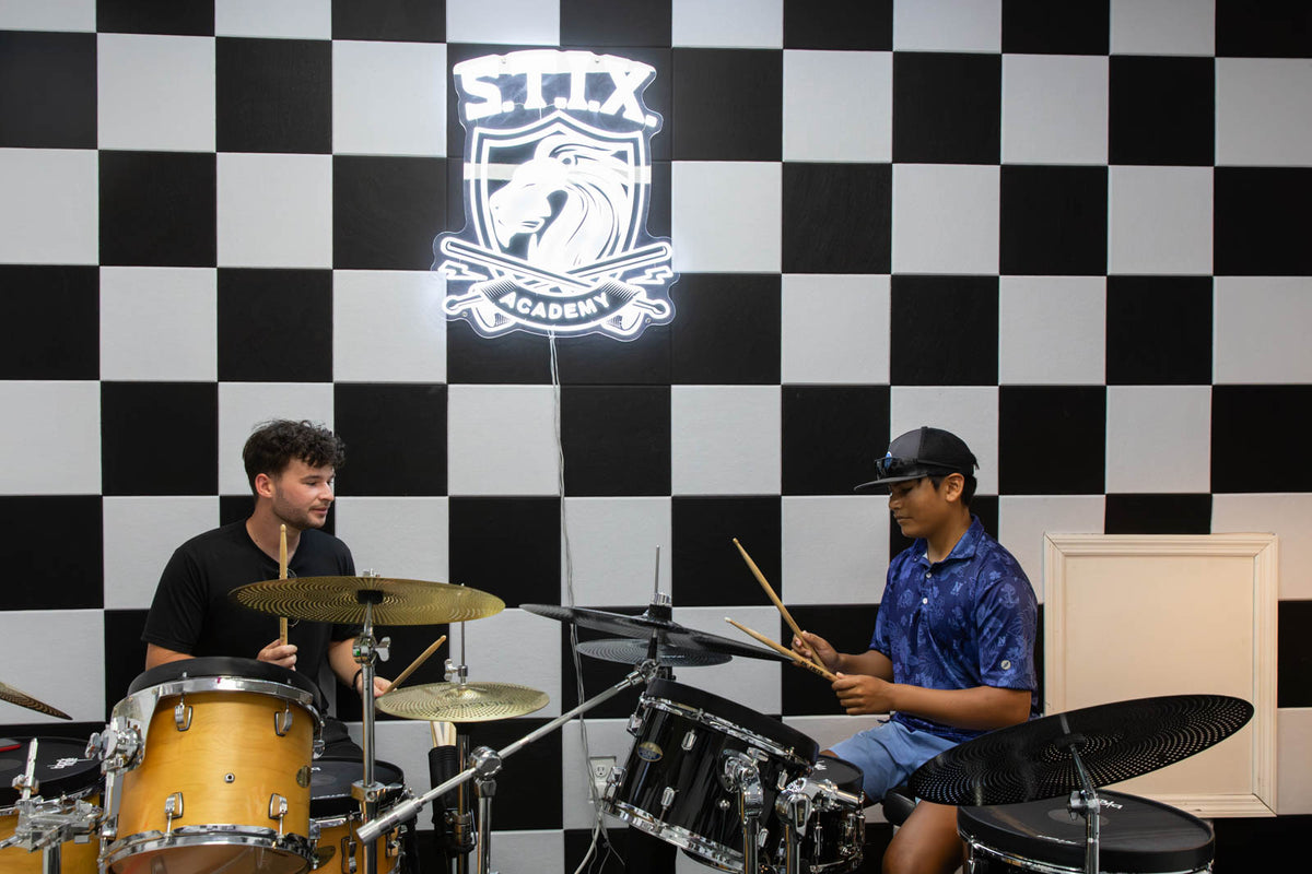 Two men playing a drum set in front of a black and white checkered wall with a glowing STIX Academy logo. One drummer is in a black shirt and the other is in a blue patterned shirt.