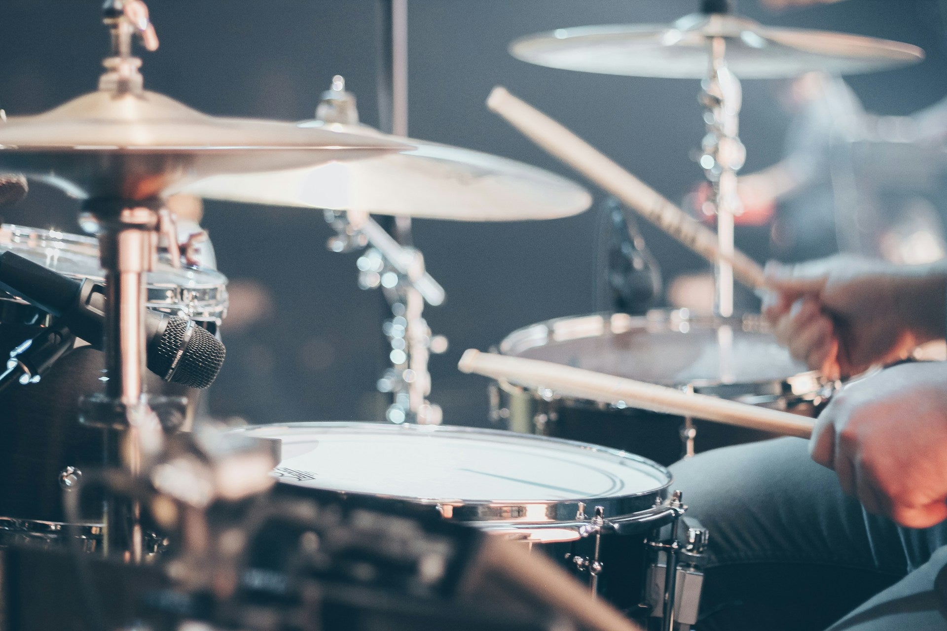 A close-up action shot of a drummer holding drumsticks in front of a drum kit with cymbals, captured in moody stage lighting with a shallow depth of field.